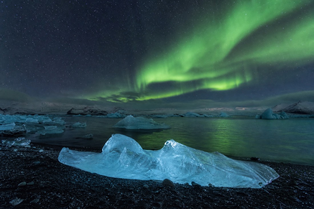Green aurora over Jökulsárlón with a glowing iceberg on the black beach as foreground on a clear winter night