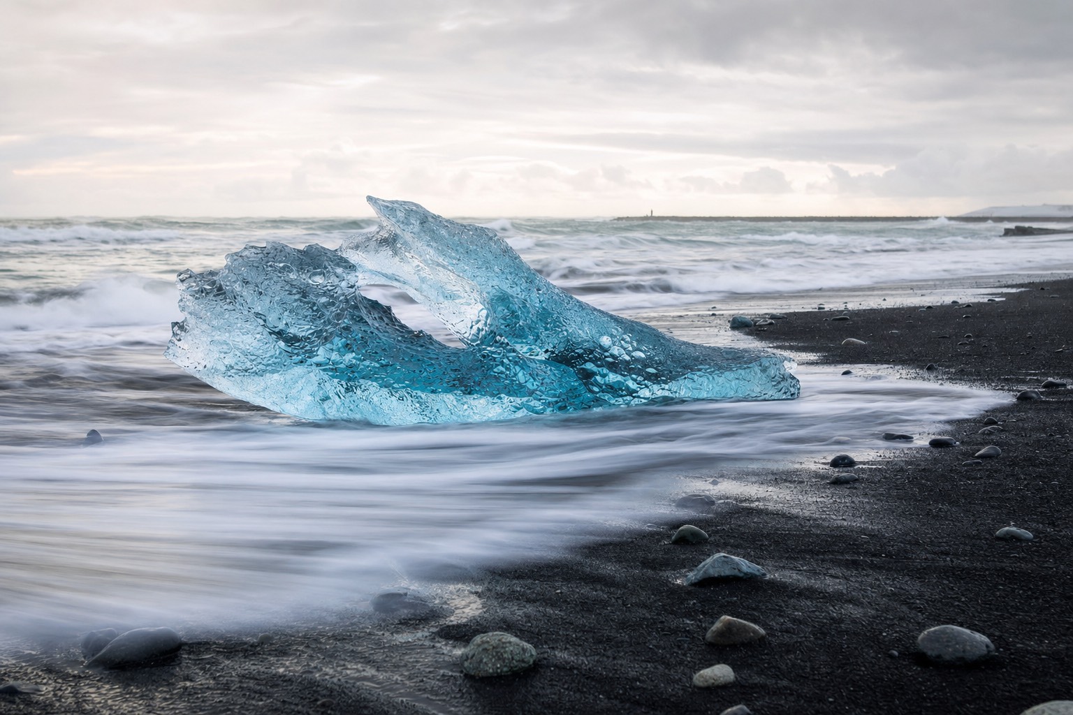 A photographer's low view along black sand at Diamond Beach with a glowing blue iceberg and surf swirling around it under soft Icelandic light