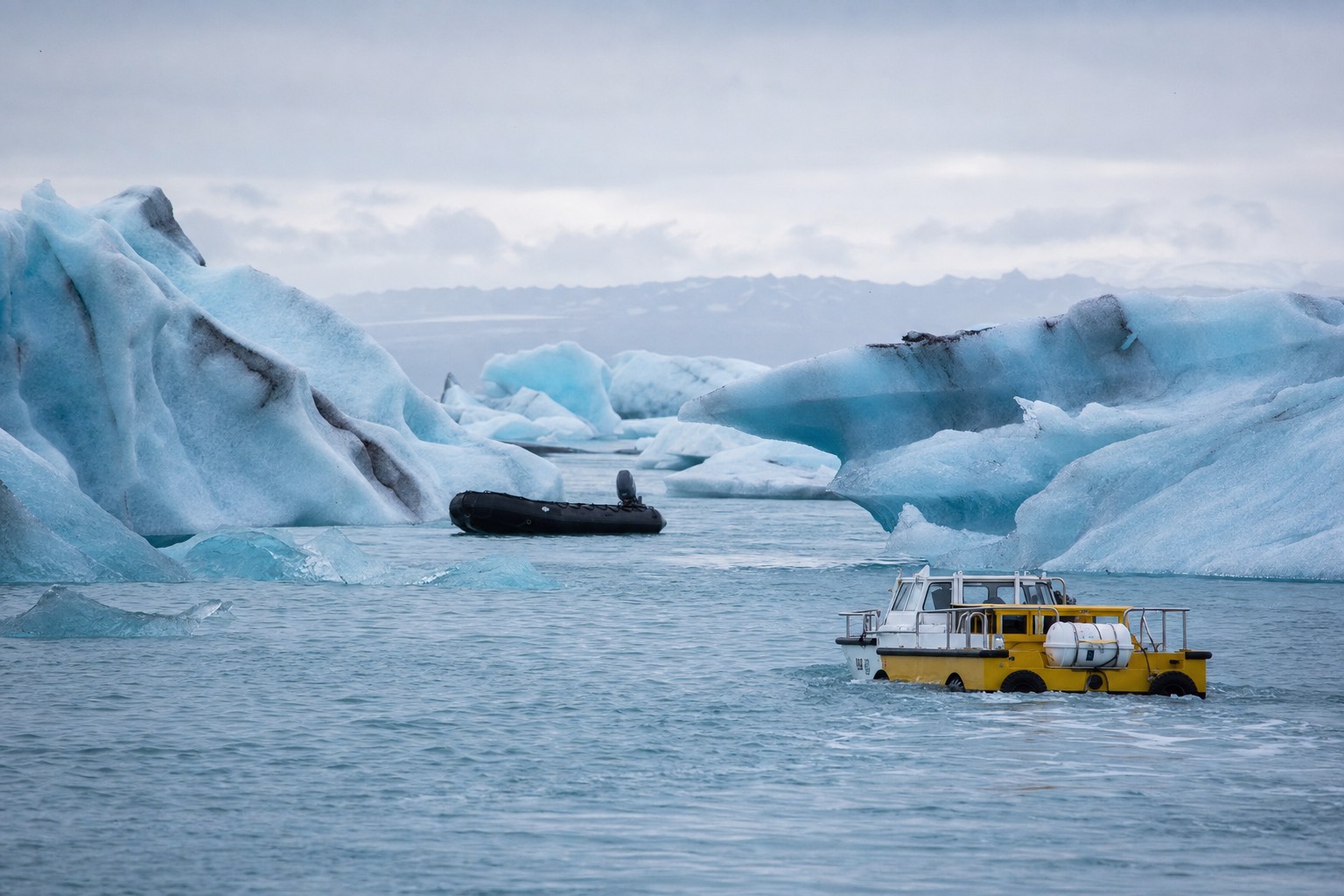 An amphibian boat and a black Zodiac inflatable threading between blue icebergs on Jökulsárlón Glacier Lagoon under low Icelandic light