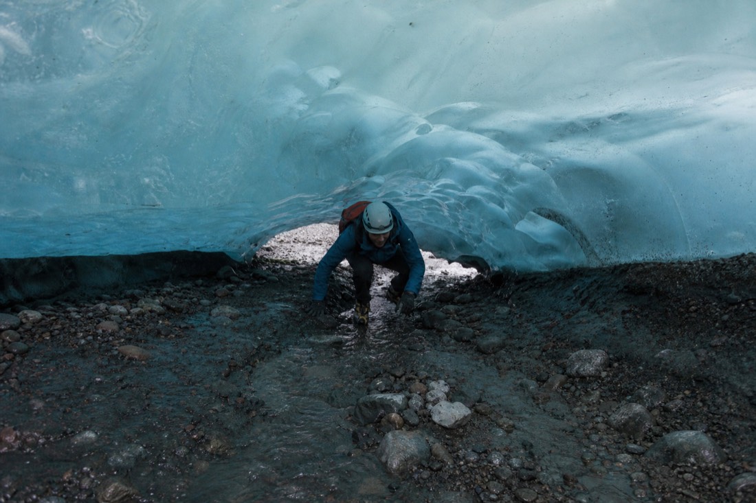 A guide in crampons and helmet leading visitors through the low entrance of a natural ice cave on Vatnajökull glacier