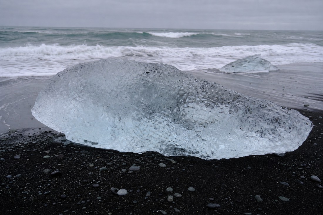 A person-sized block of clear glacier ice resting on wet black sand with waves breaking behind it at Diamond Beach