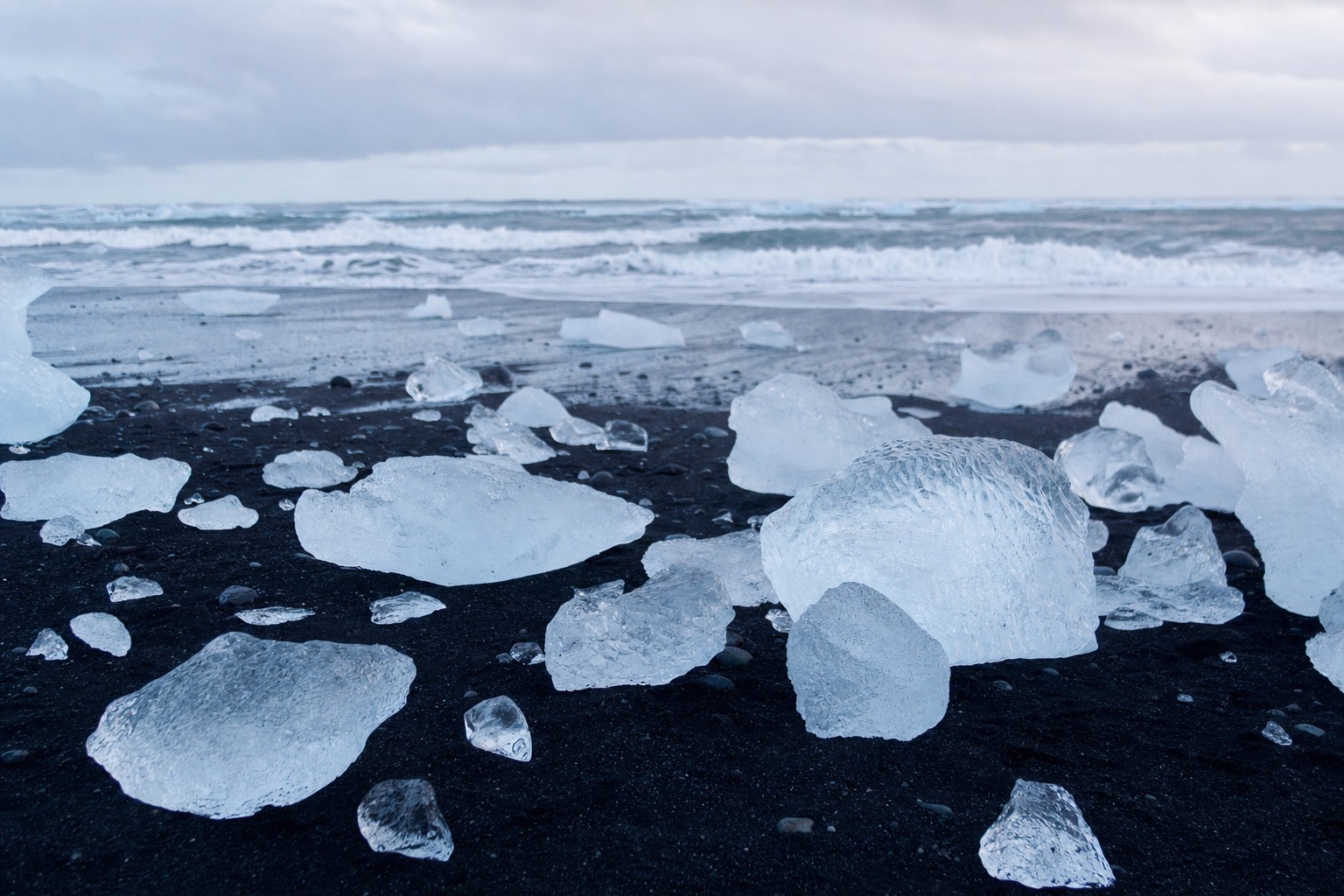 Crystal-clear and pale-blue chunks of glacier ice scattered across black volcanic sand at Diamond Beach under low Icelandic light