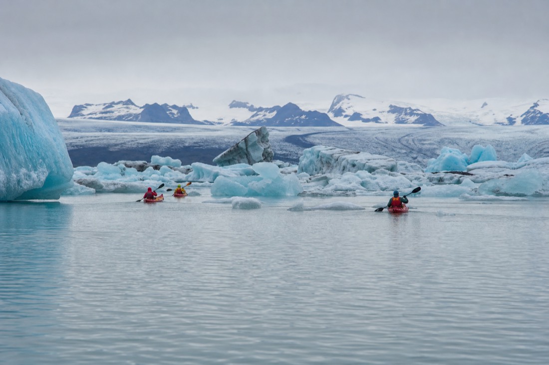 A small guided group of kayakers in drysuits paddling past blue and white icebergs on the still surface of Jökulsárlón lagoon