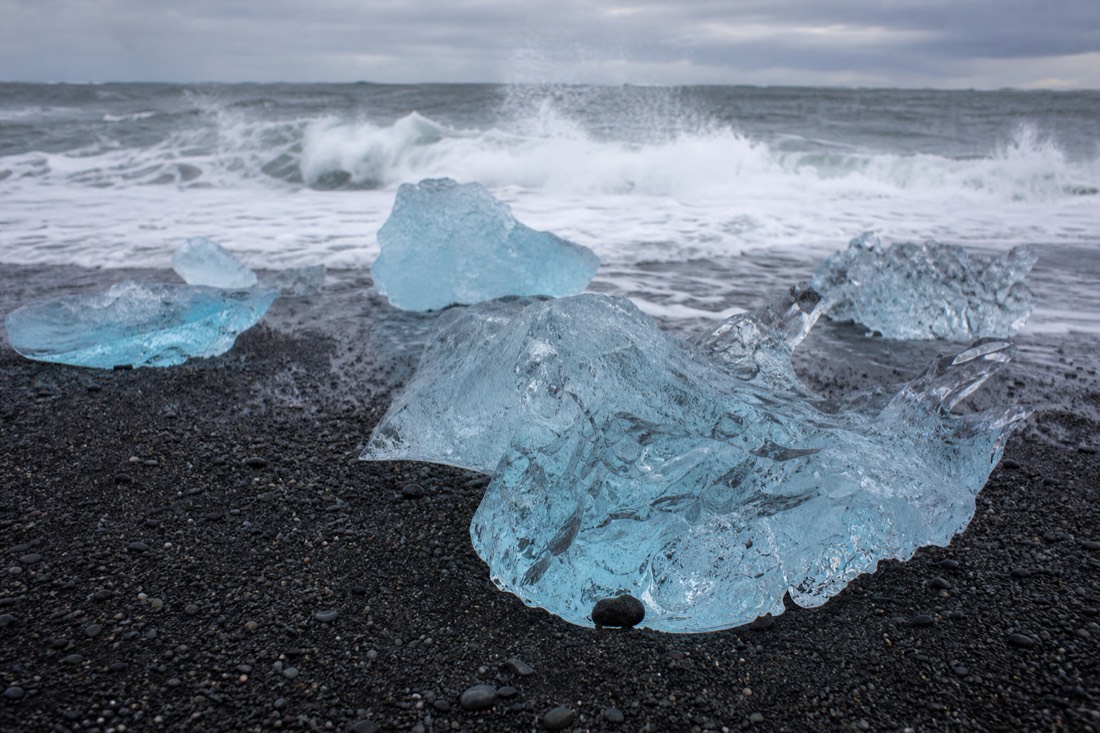 Clear and blue chunks of glacier ice resting on the black sand of Diamond Beach with waves behind