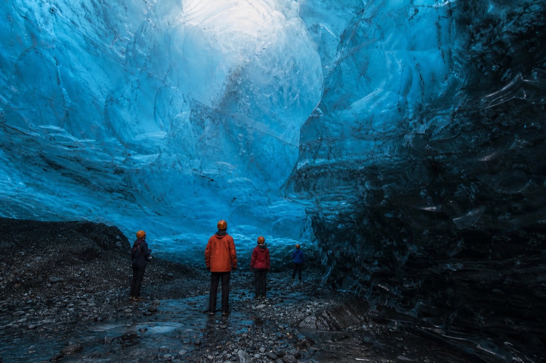 Visitors in helmets standing inside a blue natural ice cave under Vatnajökull, light filtering through the glacier ceiling