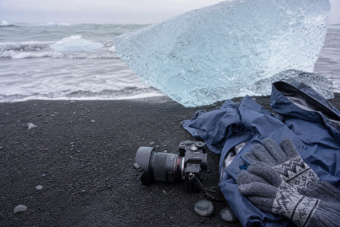 Warm gloves, a waterproof jacket and a camera resting on the black sand beside a chunk of glacier ice at Diamond Beach, Iceland