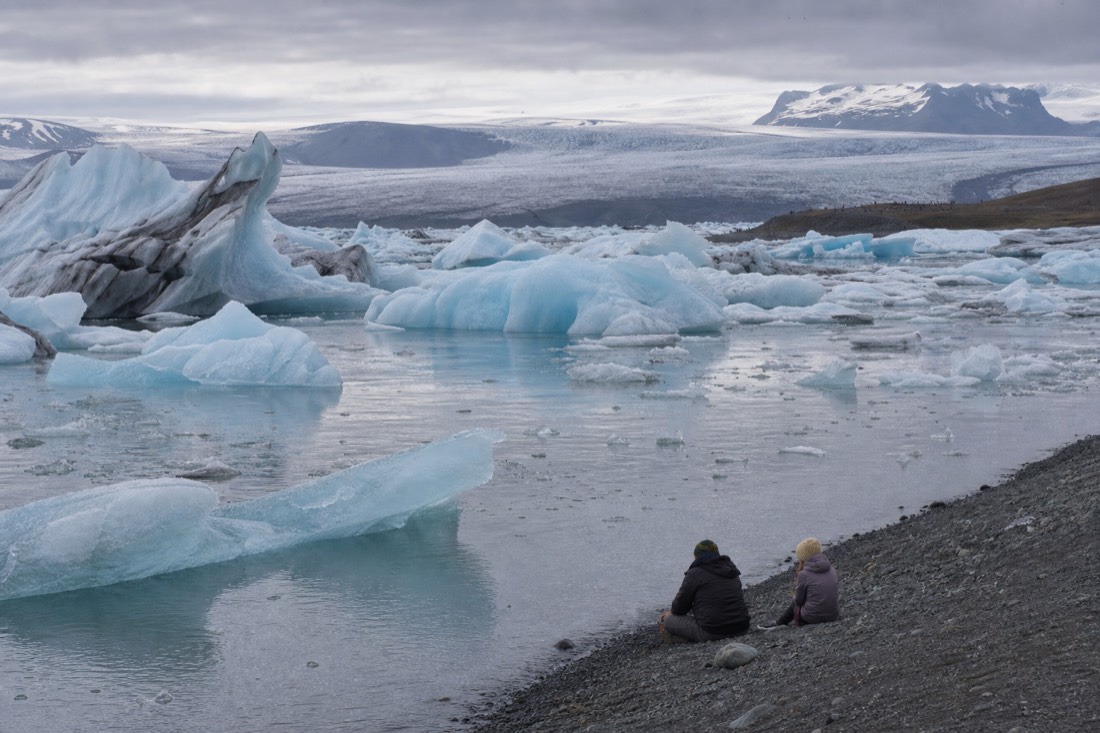 Visitors watching blue icebergs drift near the shore of Jökulsárlón Glacier Lagoon under a grey Icelandic sky